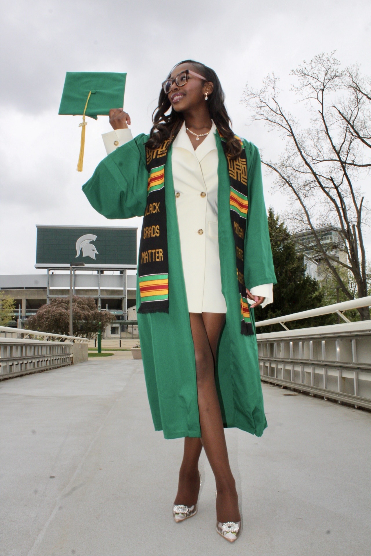 Graduate in a green commencement gown holding her cap near Spartan Stadium